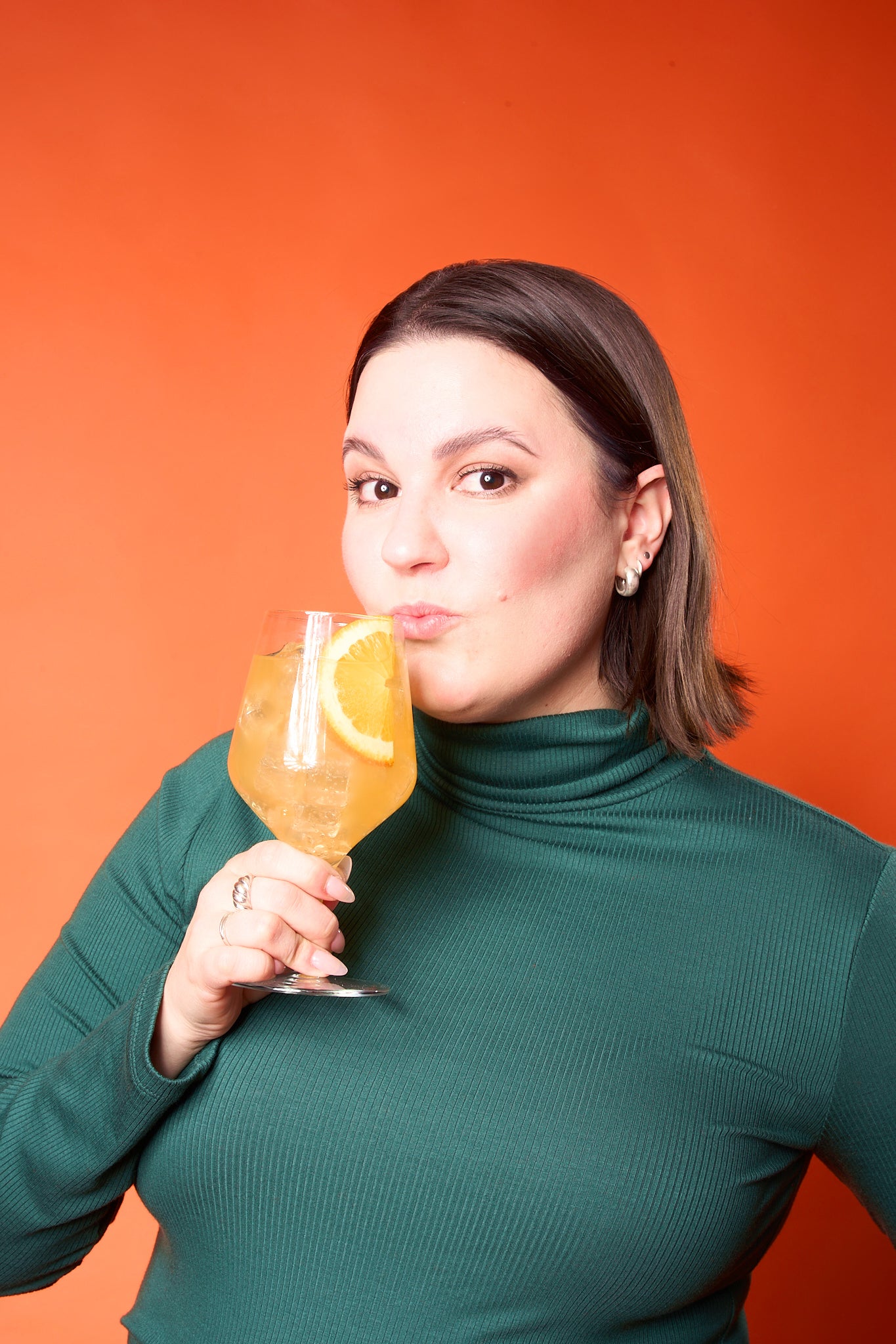 a person posing while wearing a green sweater and holding a glass of orange thc spritz liquid