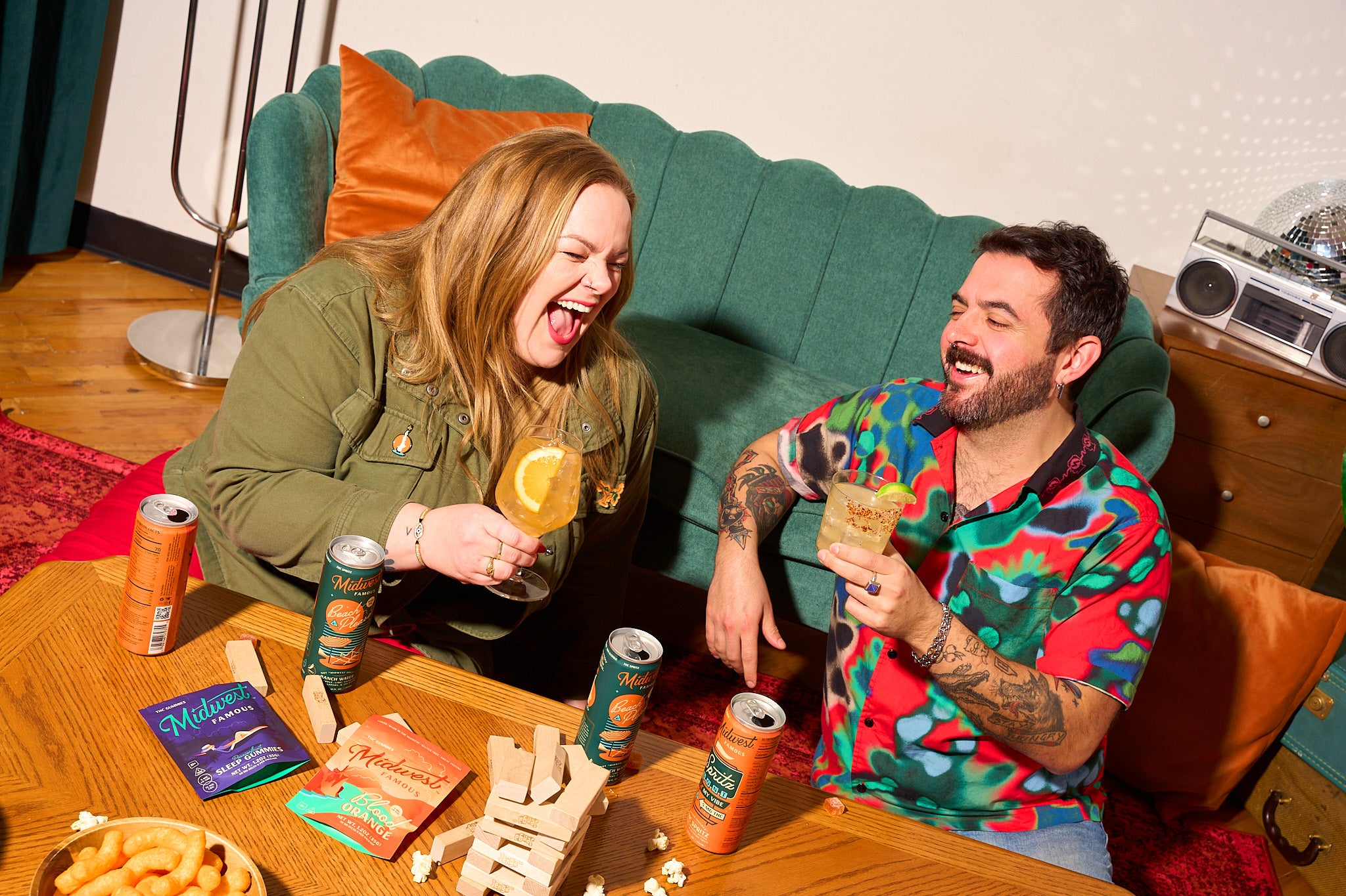 two people laughing while holding glasses of orange liquid in front of a green couch