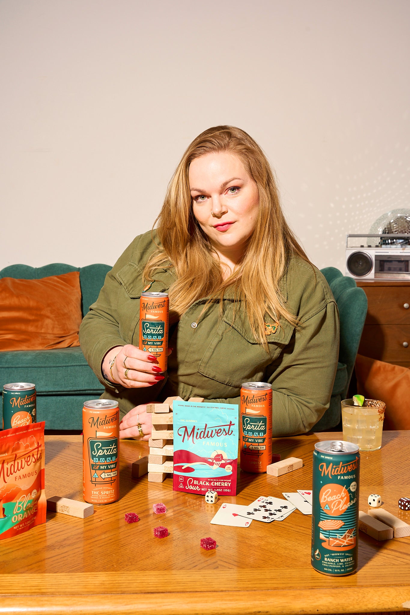 a woman holding an orange can of thc spritz with a table in front of her holding cards, gummies, and more cans of thc drinks